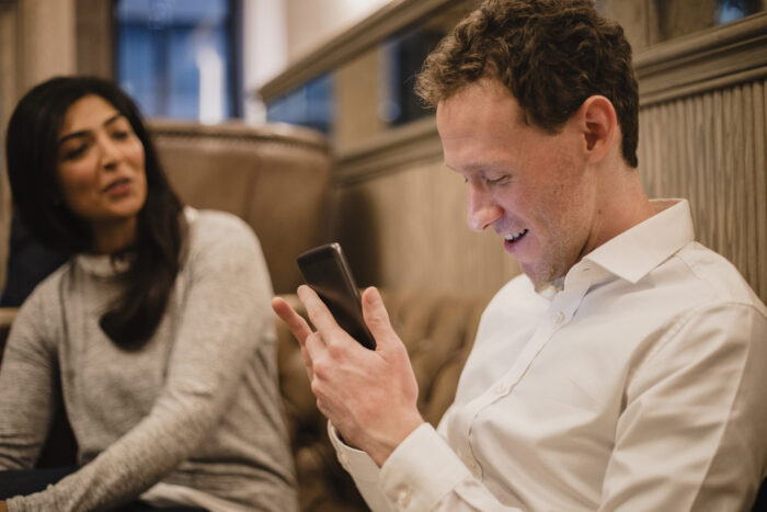 A man with his smartphone in his hand and he is using a visually impaired mobile app to help assist him, a female can be seen sitting down next to him.