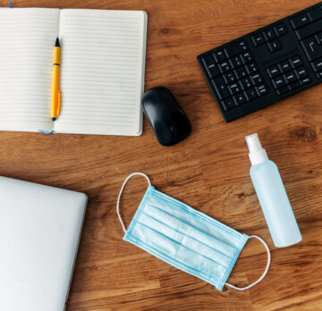 Face mask and hand sanitizer on a wooden desktop, near keyboard and notepad