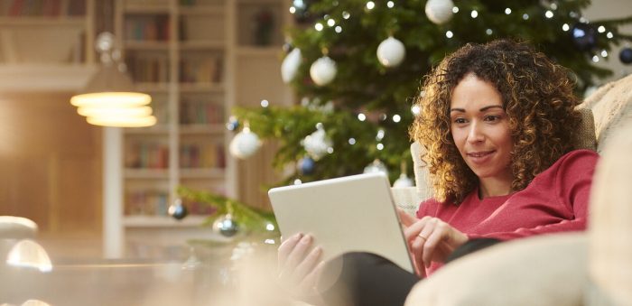 A lady sitting on a sofa, shopping on a tablet with a Christmas tree in the background