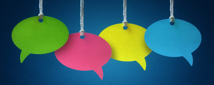 (alt=”Green, pink, yellow, blue paper speech bubbles hung by string infront of a blue background”)